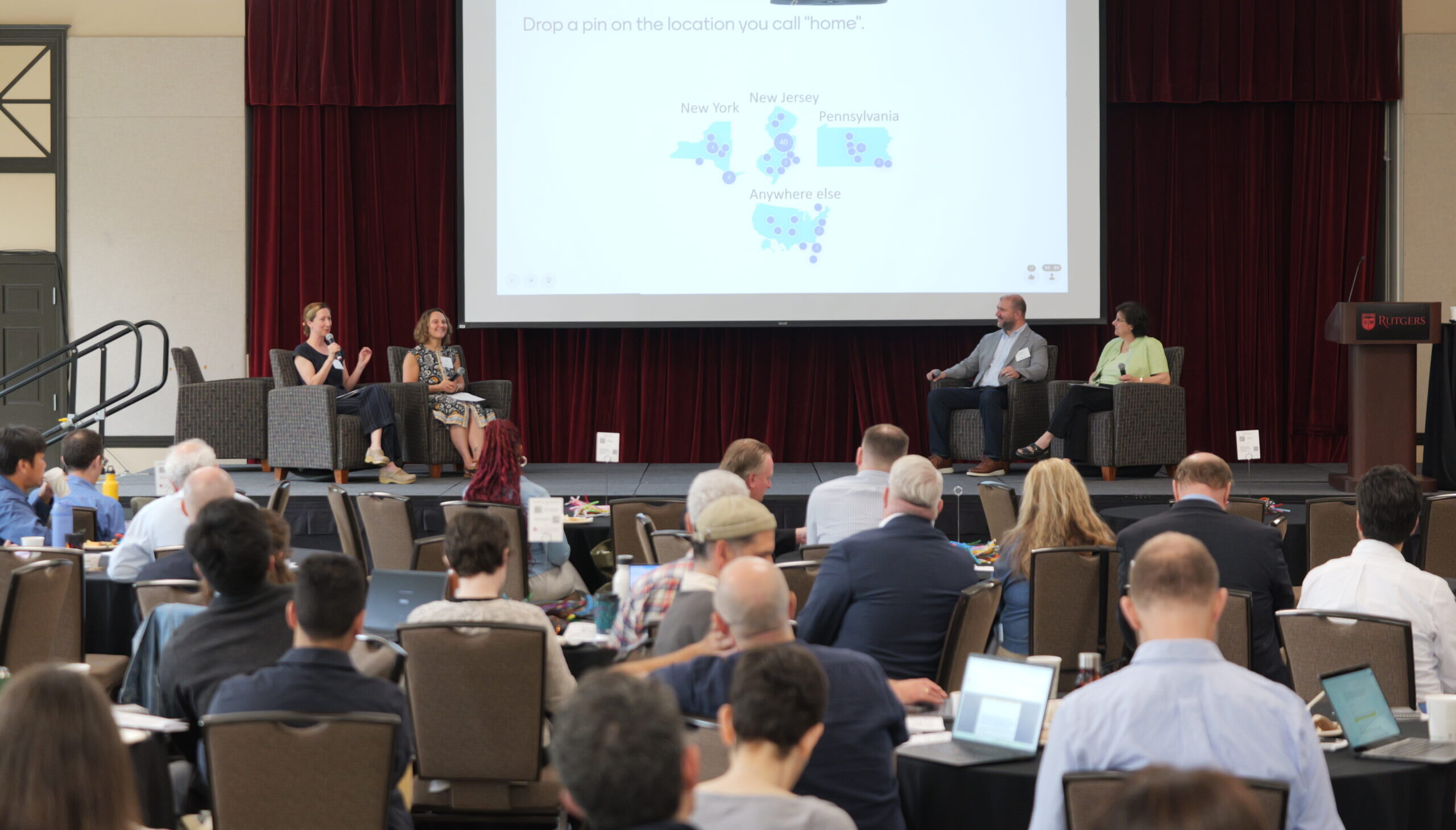 Meeting participants in audience. Panelists on stage. Projector shows promot that reads drop a pin on the location you call home.